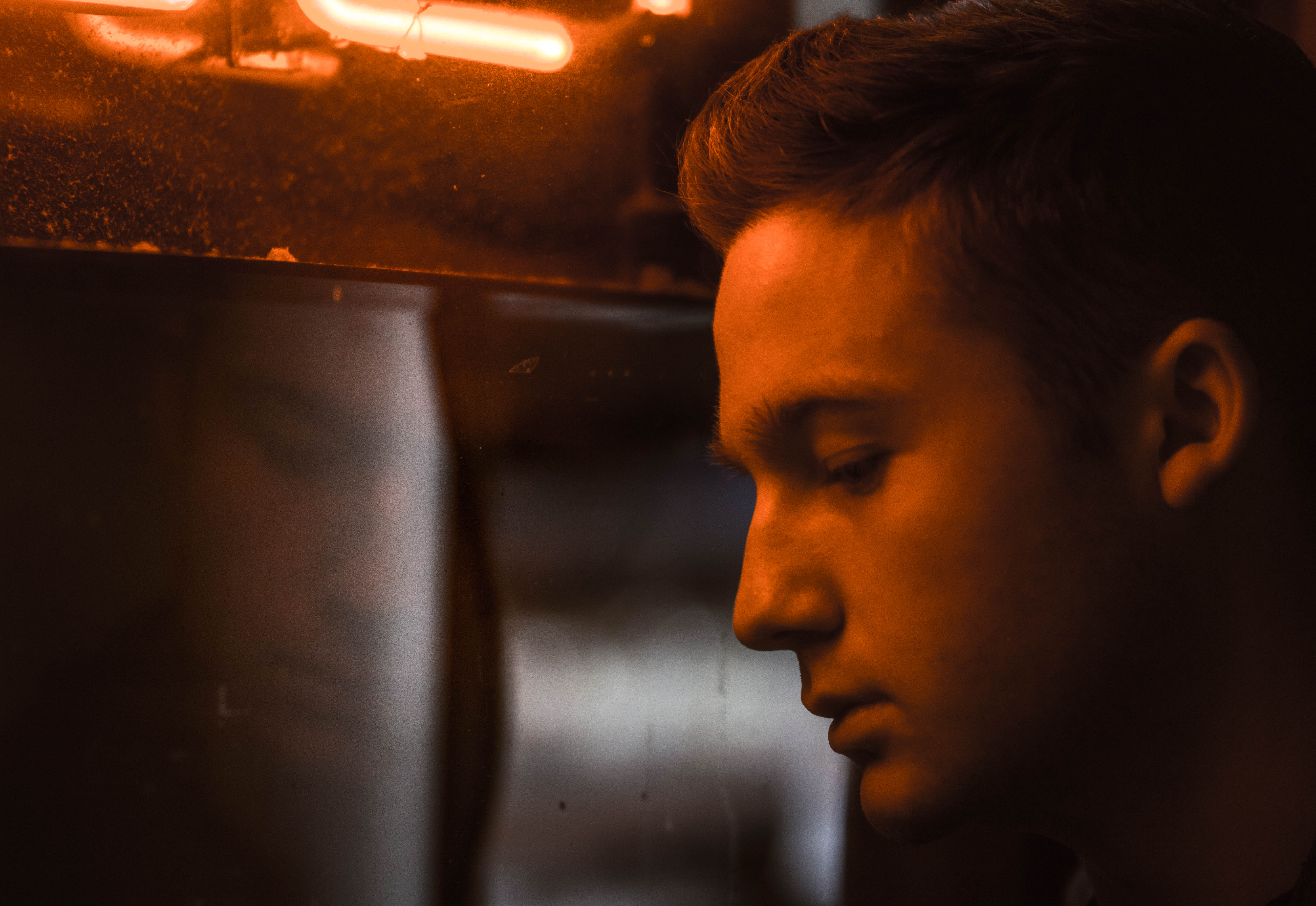 male figure stares into reflection in glass below orange neon glow, moody portrait