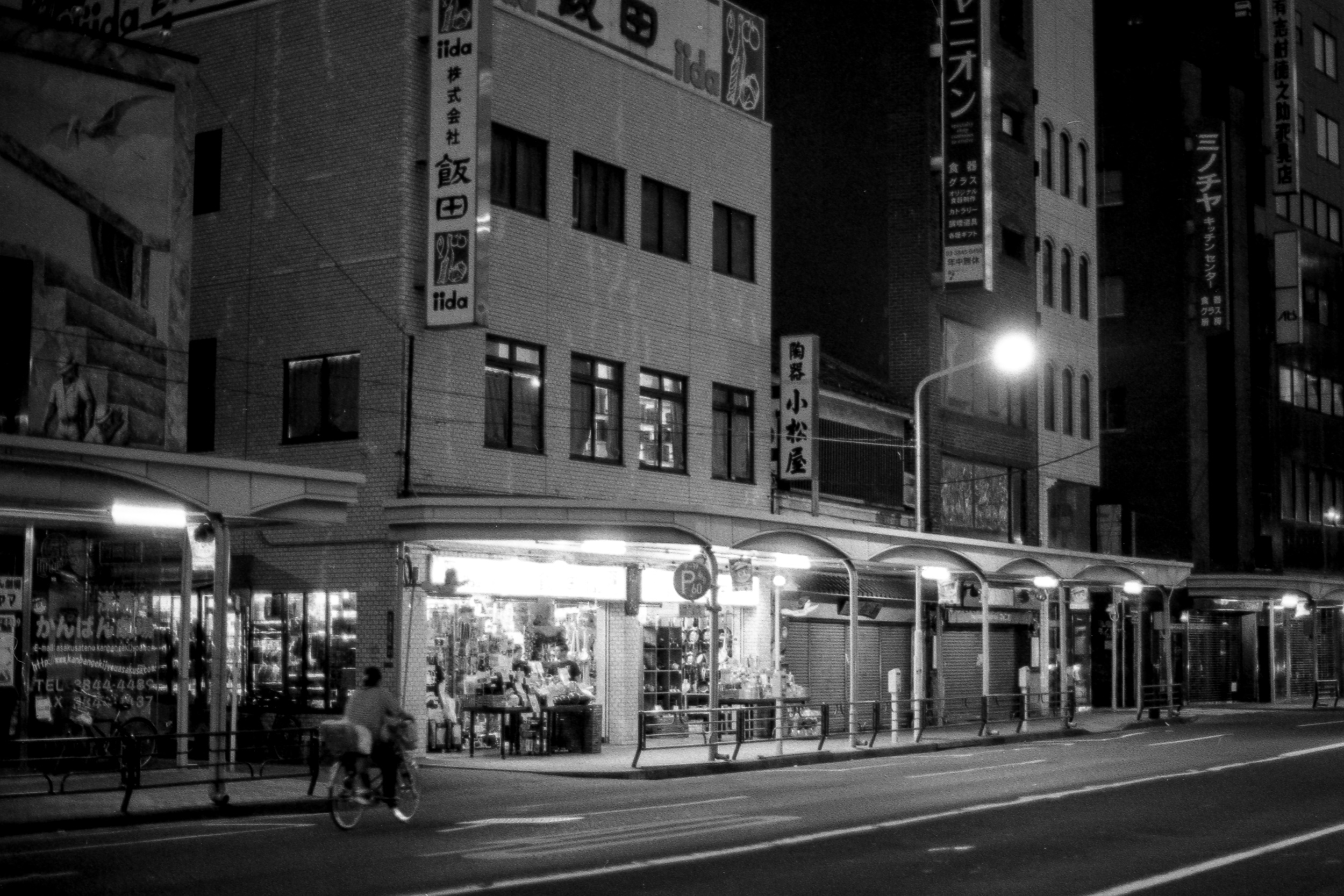 lit japanese shop on a dark street at night with person biking in front, in asakusa, 35mm black and white film