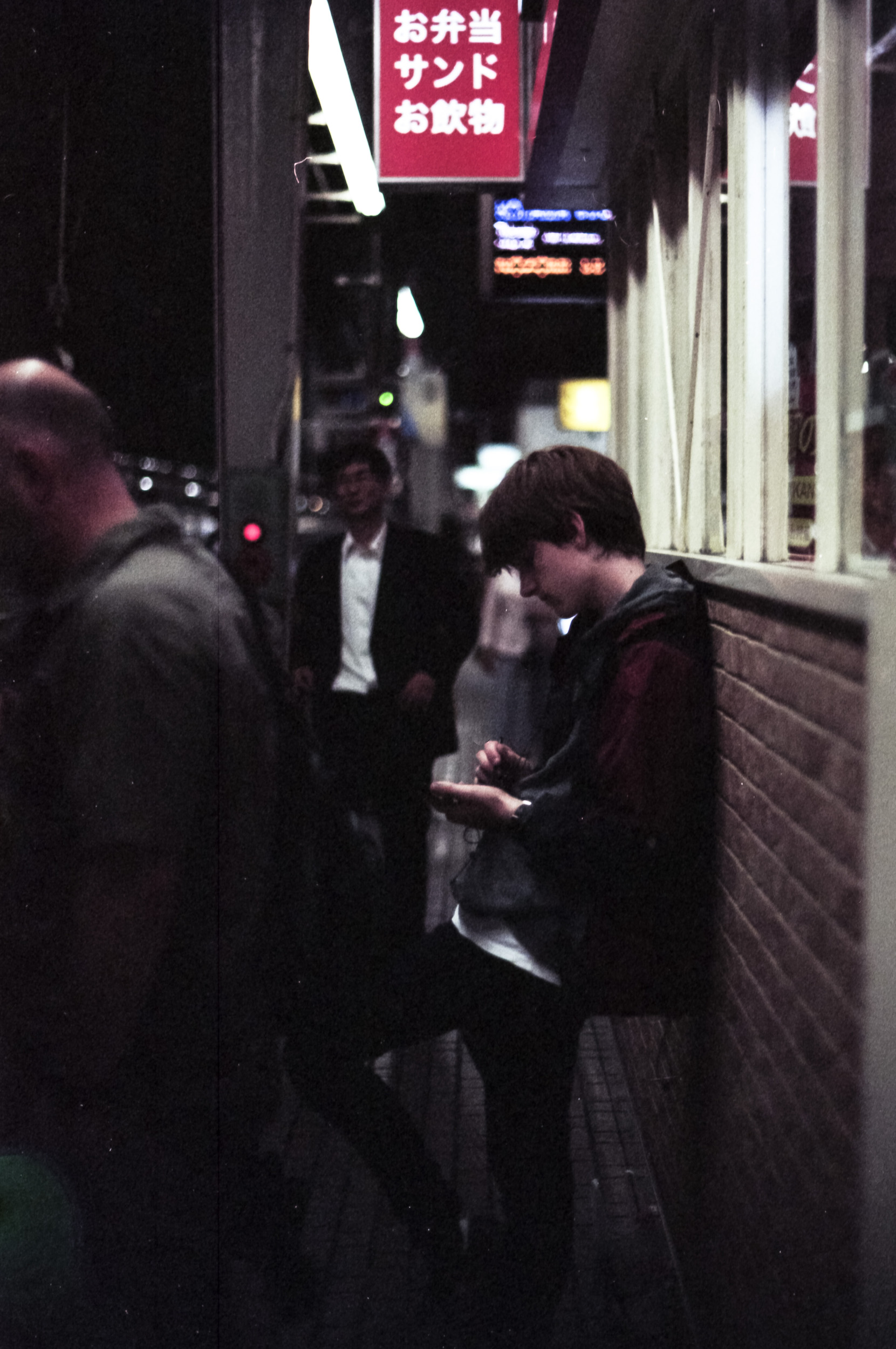 male figure leans against wall looking down at phone, japanese train station at night, 35mm film