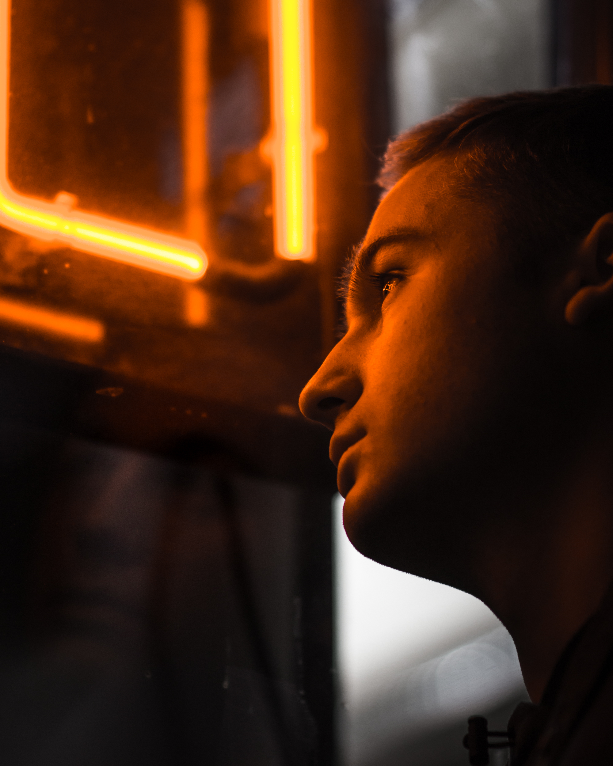 moody portrait of somber male looking down in front of orange neon sign with his face reflected on glass window pane, night