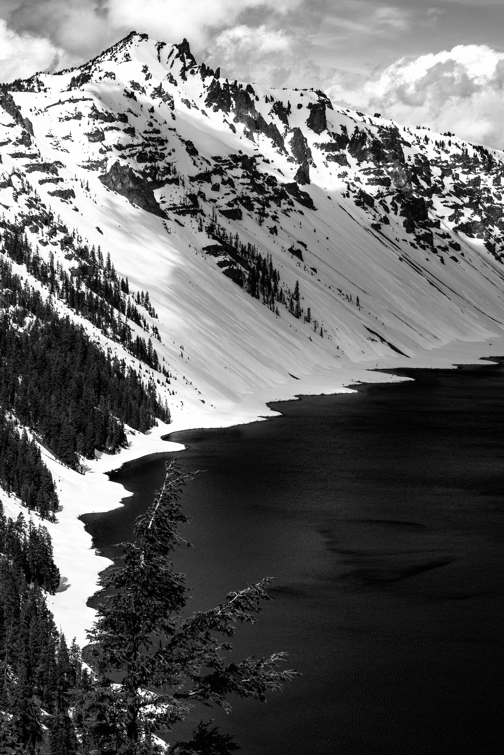 high contrast black and white photo of crater lake with snowy hills on a partly cloudy day