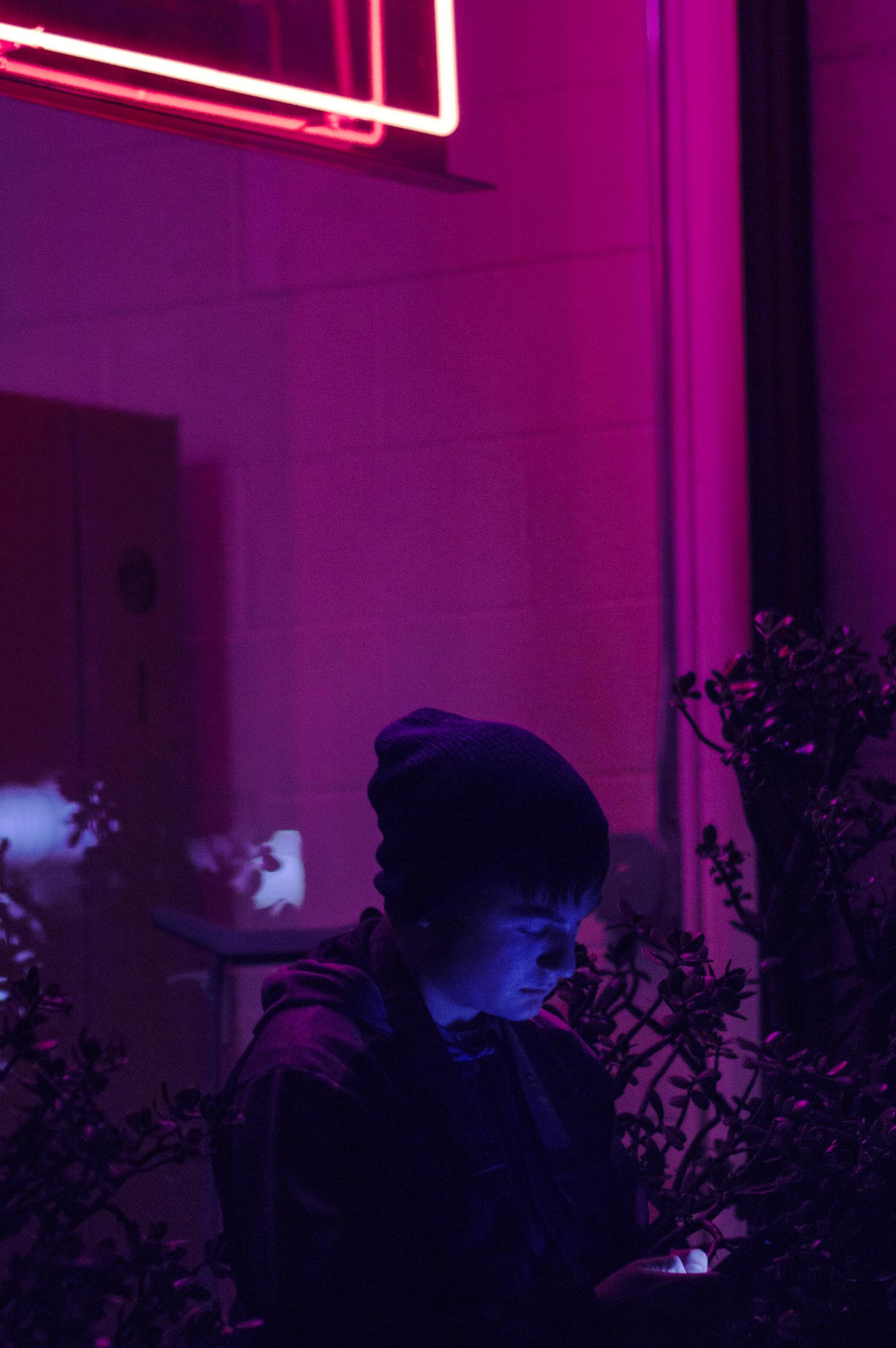 male figure wearing beanie browses phone beneath shopfront window with neon lights