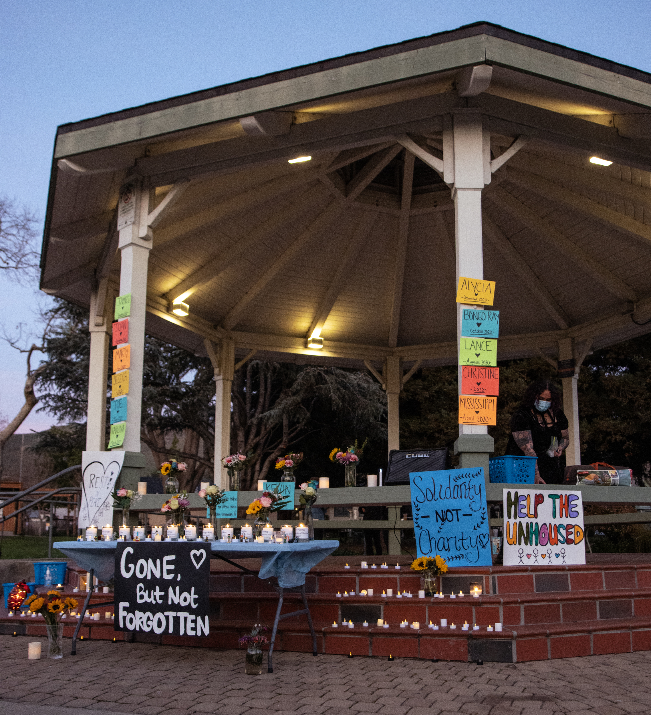 a decorated gazebo at twilight with candles for a vigil and signs promoting support for the unhoused