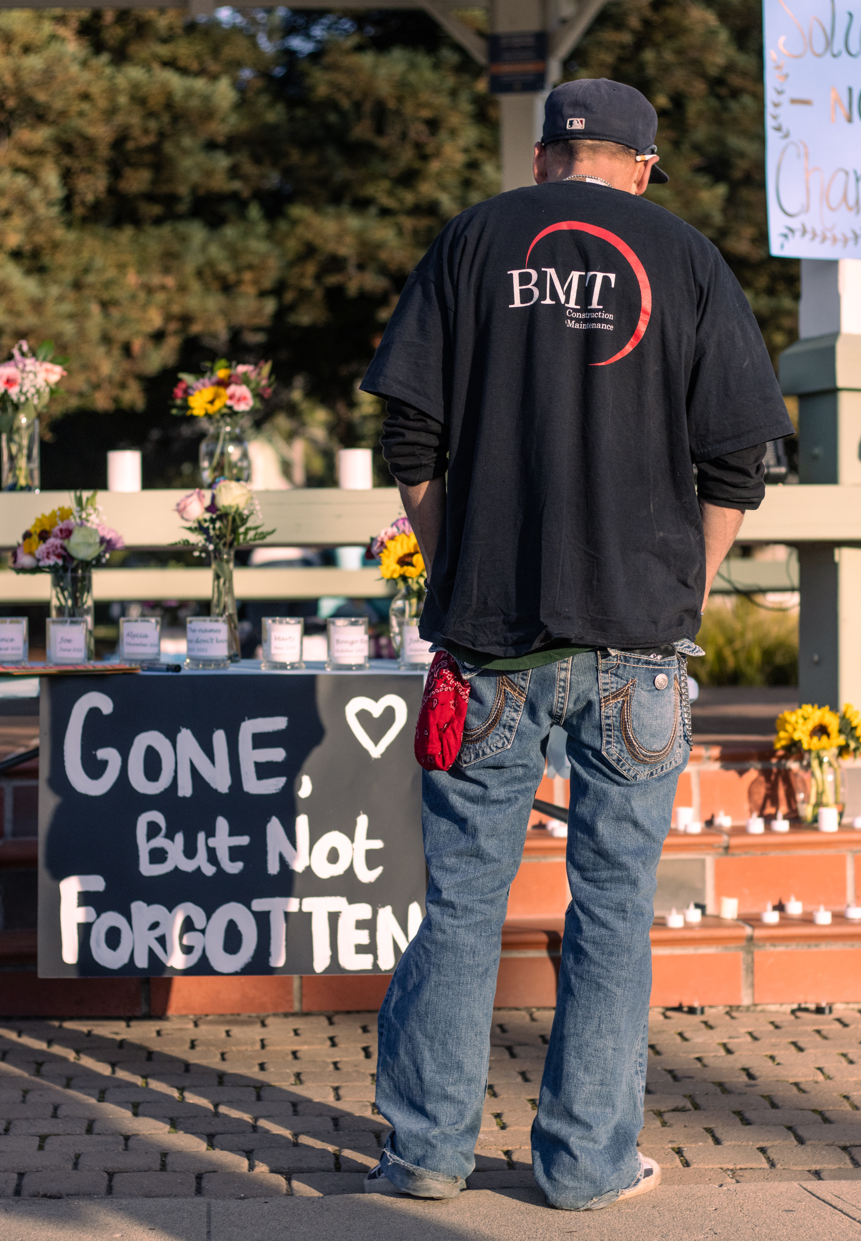 a middle aged man faces away from the camera, observing a sign which reads gone but not forgotten