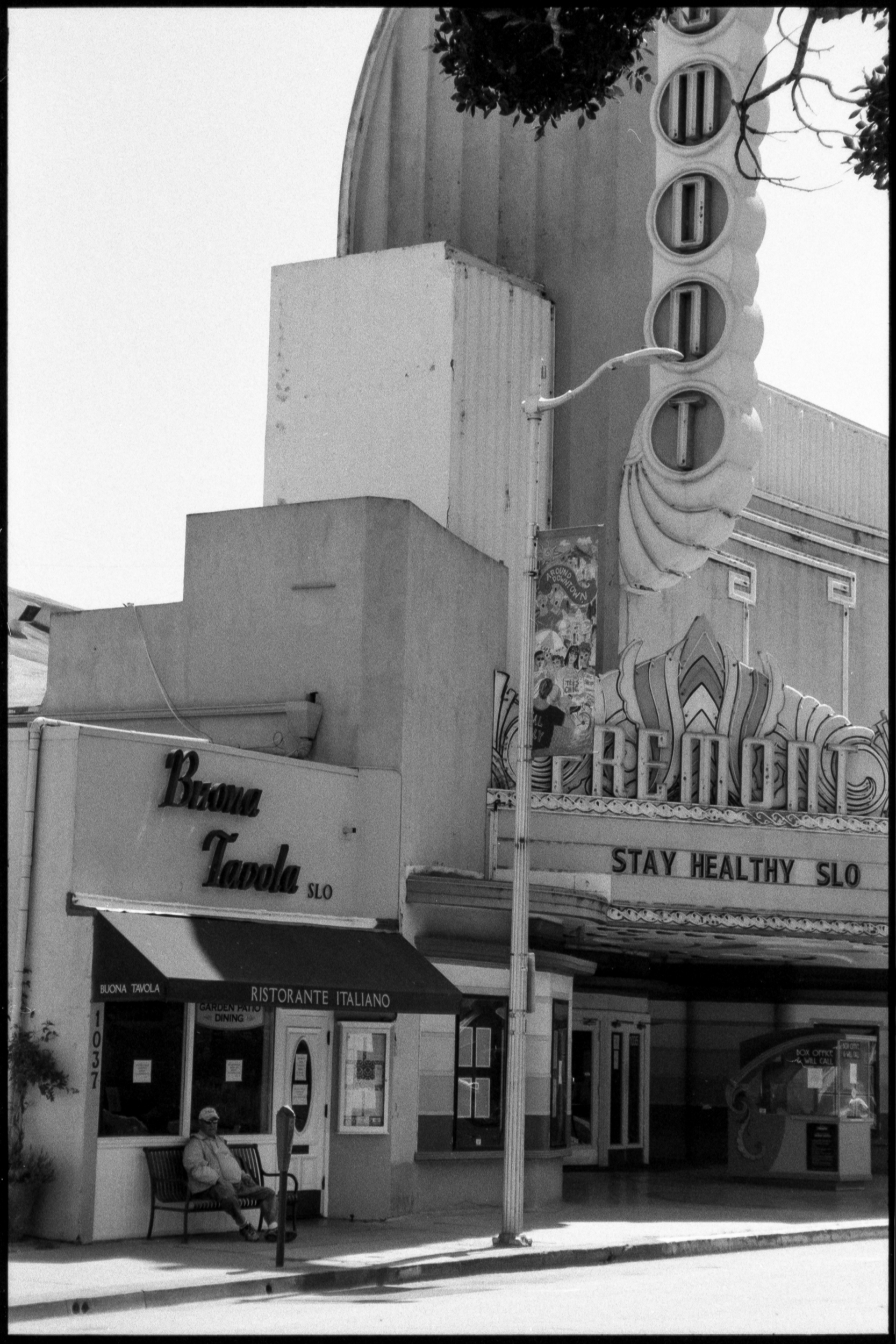 a single middle-aged man sitting alone in front of an italian restaurant and the fremont theater with a matinee which reads stay healthy slo
