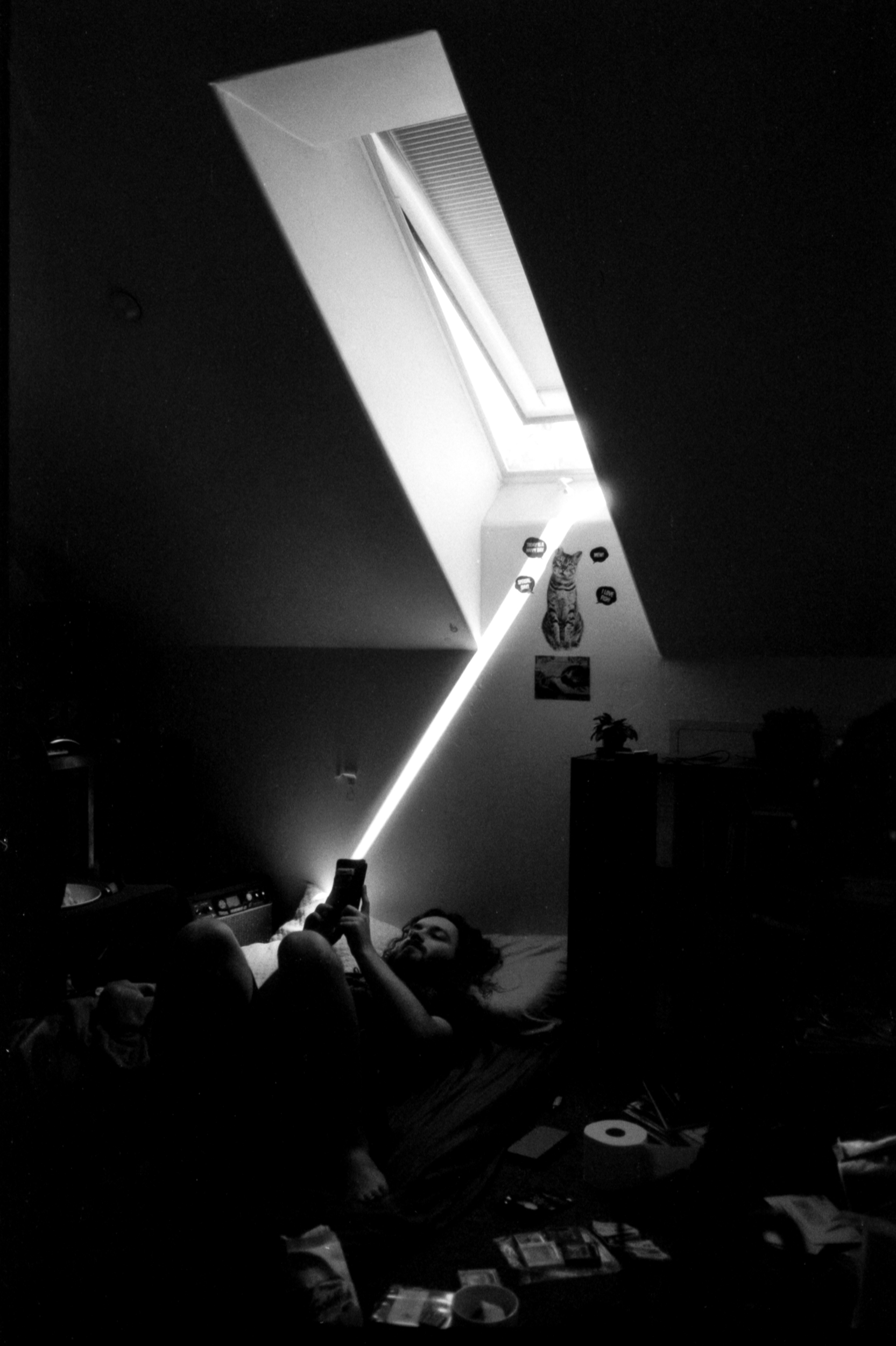 a male figure lying in bed in a dark cluttered room with a rectangular skylight directly above casting a harsh linear ray of light onto his outstretched hand holding a phone