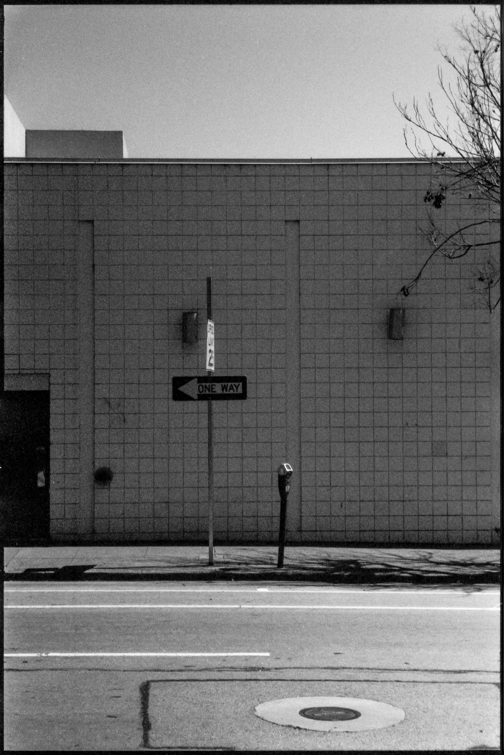 a one way sign next to a parking meter on an empty street with a grid-tiled building behind