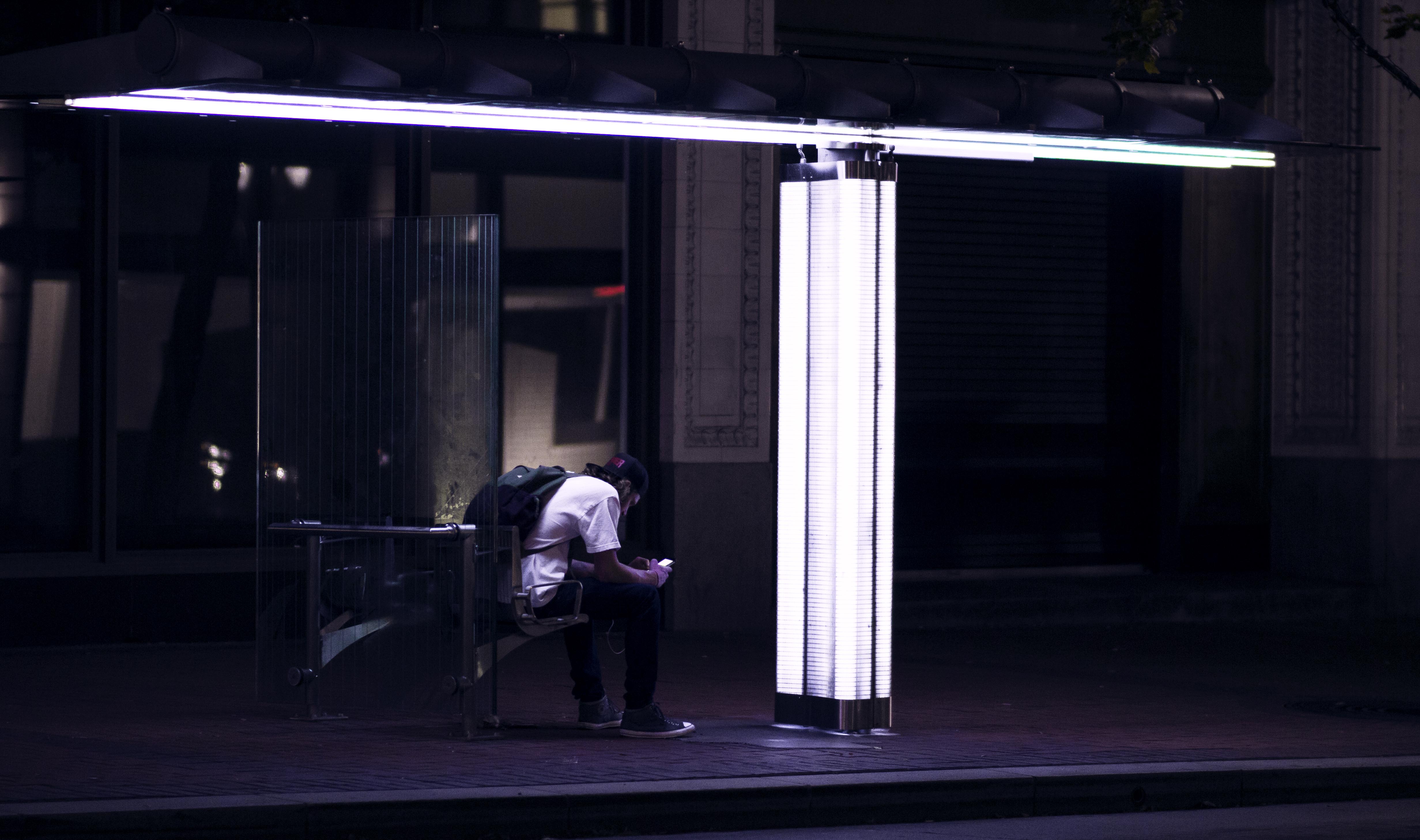 man looking at phone sitting at cantilever lit bus stop at night, portland oregon